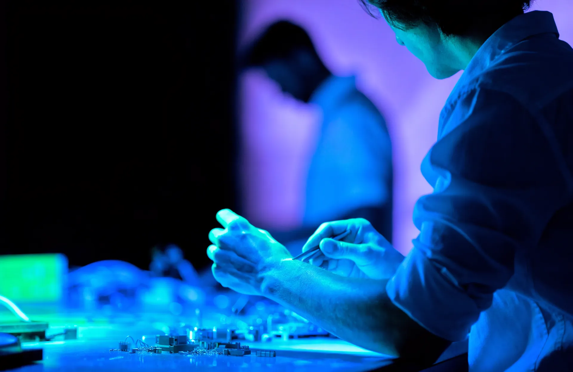 Engineer at a workbench assembling hardware under blue task lighting, seen from the side